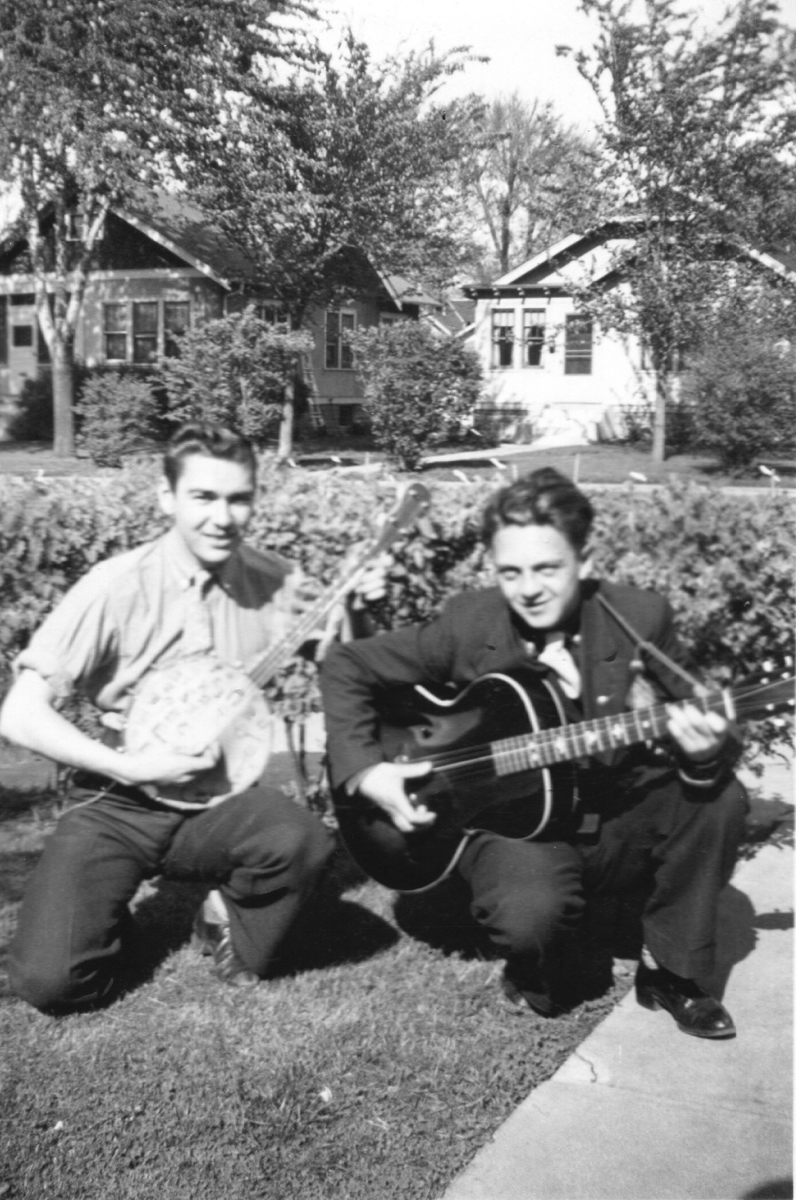 two late teen boys dressed up holding a banjo and a guitar on the front lawn of a house