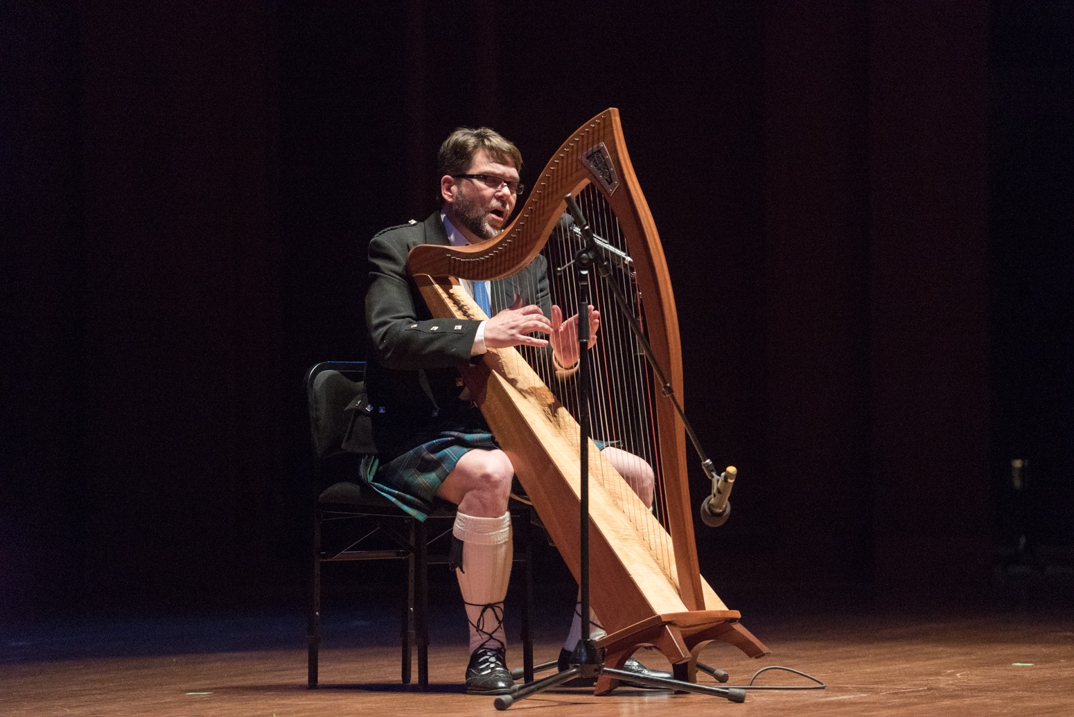 man in kilt seated behind harp on stage singing