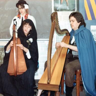 two high school students in costumes seated playing the harp