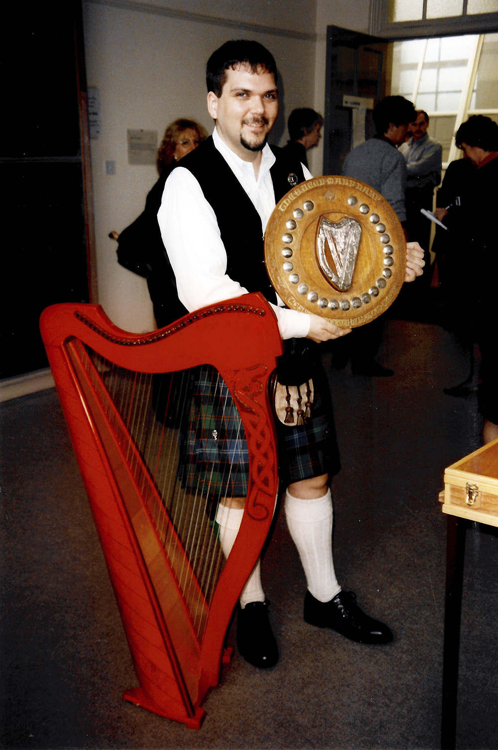 man standing next to red harp carrying large round trophy